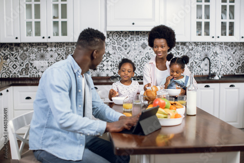 African american woman feeding two pretty daughters with breakfast while husband working on digital tablet. Happy family sitting on bright kitchen during morning time.