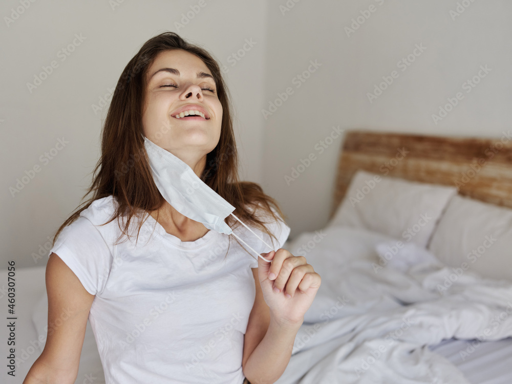 a woman with a medical mask in her hand sits on a bed In a bright room tilt her head back a woman with a medical mask in her hand sits on a bed In a bright room tilt her head back
