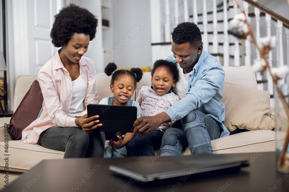 Happy african parents sitting on couch with two pretty daughters and using digital tablet for watching cartoons. Concept of family, leisure time and modern gadgets. Happy african parents sitting on couch with two pretty daughters and using digital tablet for watching cartoons. Concept of family, leisure time and modern gadgets.