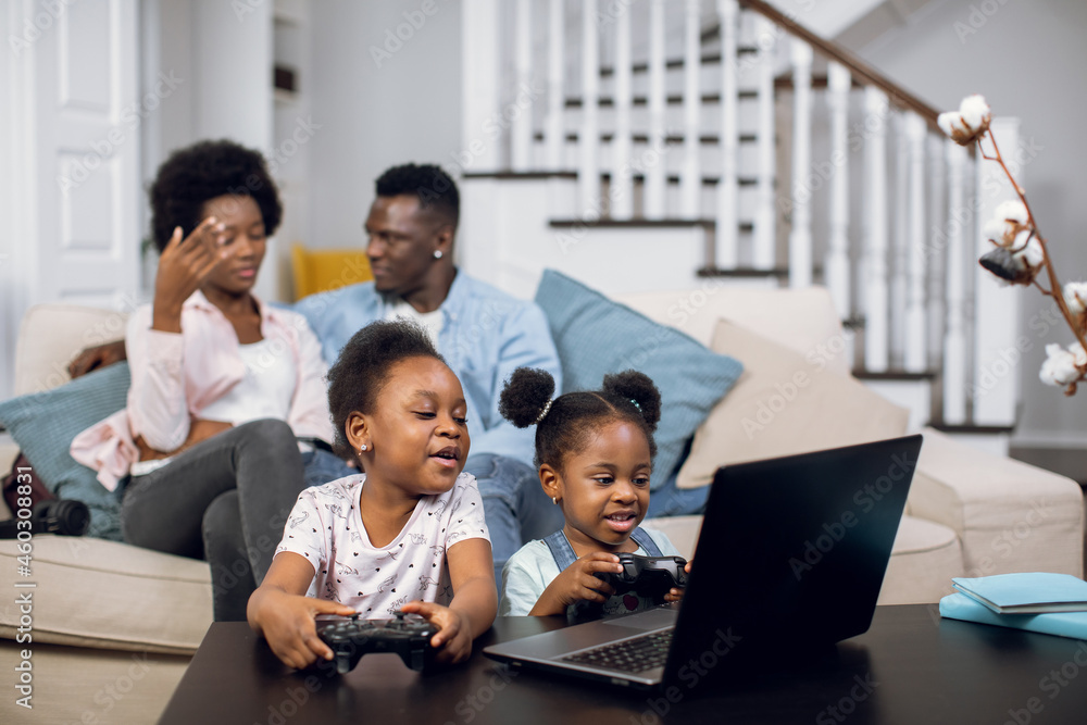 African american parents talking and hugging on couch while their cute daughters playing video games on laptop. Happy family enjoying free time at home.