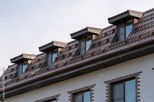 Sloping roof with metal coating of residential building with windows. Roof with drains, slopes, snow retainers, tides and dormer windows against sky