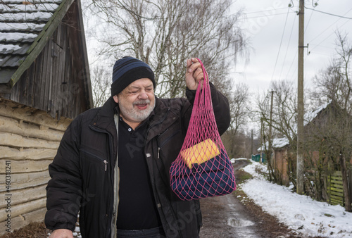 Portrait of smiling elderly peasant walking with string bag  to local bath-house on an empty street of rural village Pidstavky, Sumskaya oblast, Ukraine