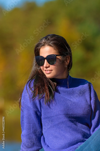 Closeup portrait of a young smiling brunette woman in a blue knitted sweater wearing sunglasses on a sunny autumn day. Colourful forest in the background.