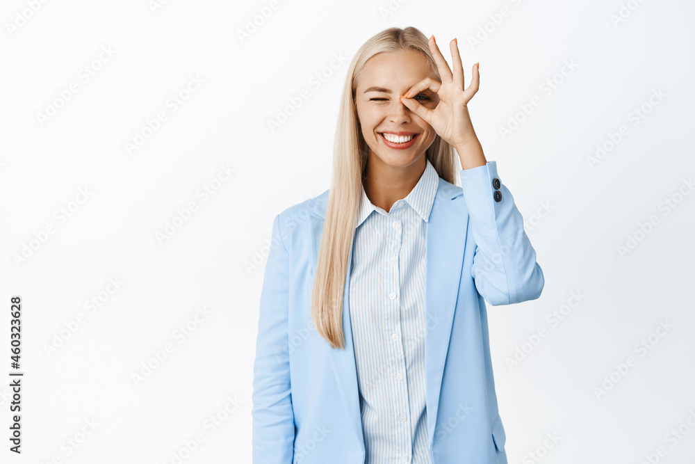Enthusiastic businesswoman showing zero, okay gesture on eye and smiling, recommending something good, standing in suit over white background Enthusiastic businesswoman showing zero, okay gesture on eye and smiling, recommending something good, standing in suit over white background