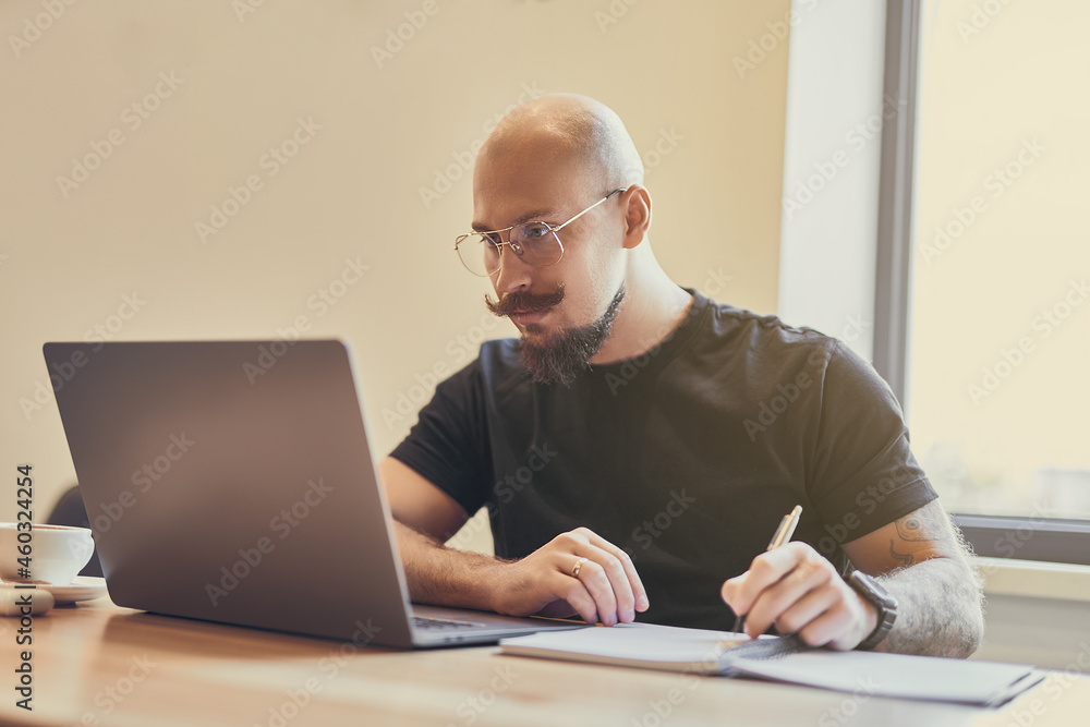 Young bald man working on laptop sitting at desk studying doing something notes. Distance Learning Young bald man working on laptop sitting at desk studying doing something notes. Distance Learning