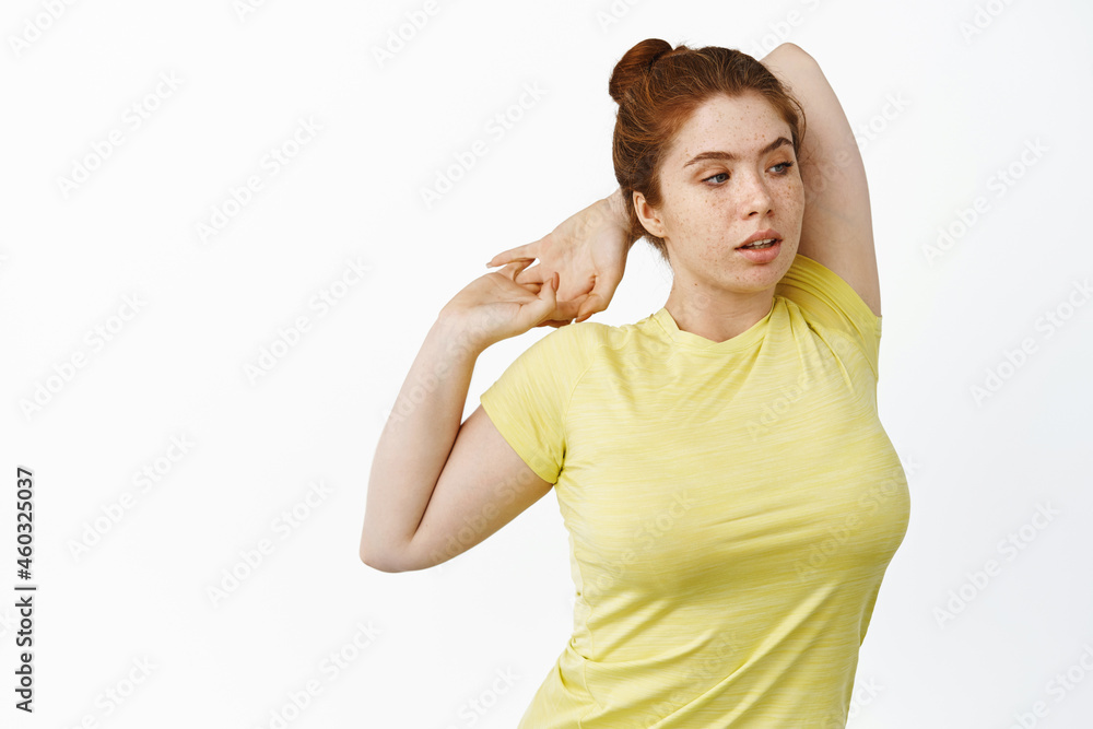 Portrait of redhead plus size woman stretching arm behind back, warm-up fitness exercises in gym, standing over white background Portrait of redhead plus size woman stretching arm behind back, warm-up fitness exercises in gym, standing over white background