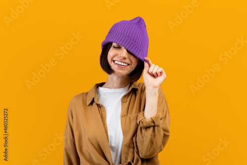 Happy young woman pulling down trendy purple hat, closing one eye and smiling broadly, orange background