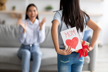 Mother's day gift. Girl congratulating excited mother with holiday, hiding greeting card and present box behind her back
