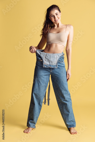 Portrait of young beautiful slim woman in blue jeans jumpersuit posing isolated over yellow studio background. Weight Loss concept.