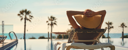 Canvas Print Rear view woman wear hat lying on deckchair near pool, put hands behind head relaxing, take sun bath, sea palm tree empty swimming pool scenery on background