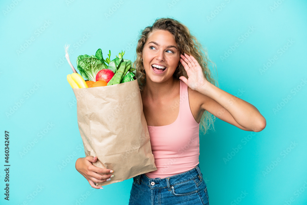 Girl with curly hair holding a grocery shopping bag isolated on green background listening to something by putting hand on the ear Girl with curly hair holding a grocery shopping bag isolated on green background listening to something by putting hand on the ear