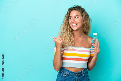 Young blonde woman with a bottle of water isolated on blue background pointing to the side to present a product