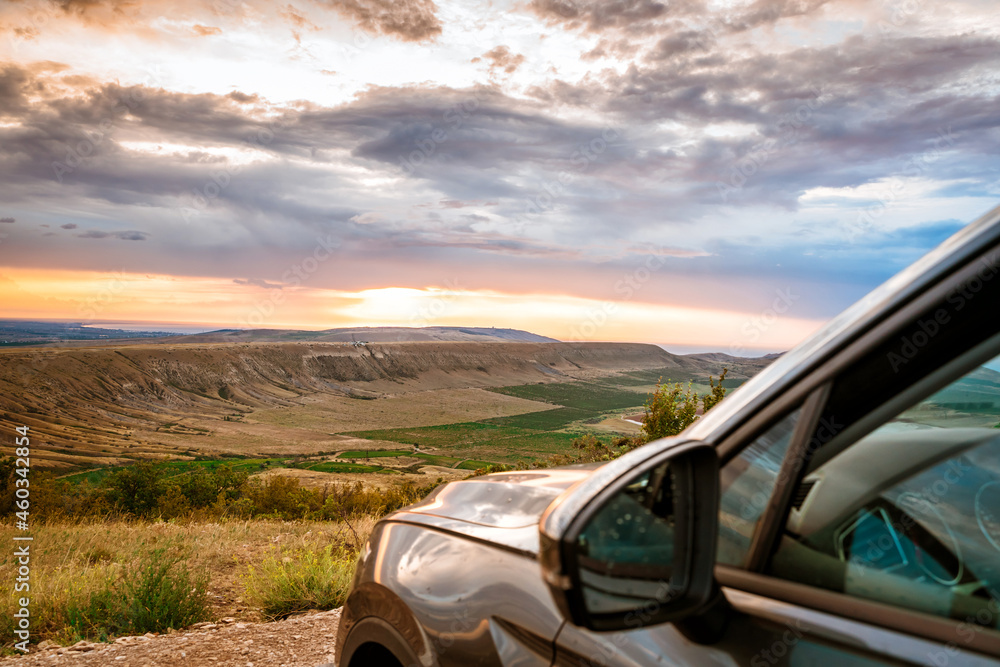 The view of the car stands in front of a picturesque mountain landscape at dawn with a dramatic sky. Colorful travel background.