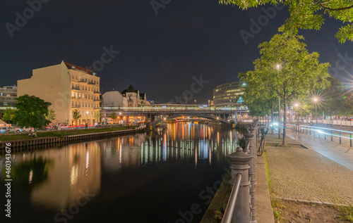 Tree lined boulevards along sides of Spree River with urban night lights reflected in calm water below railway bridge.