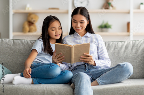 Family leisure activity concept. Young asian mother and daughter reading book together, sitting on sofa, free space
