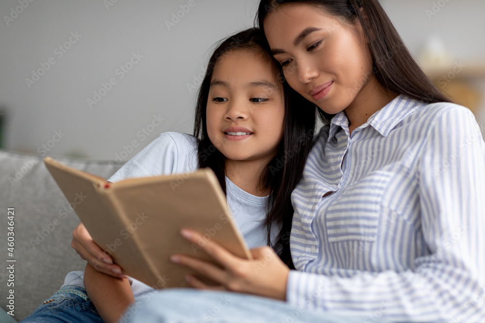Happy asian girl reading book with her mother, sitting together on sofa and smiling, spending time at home interior