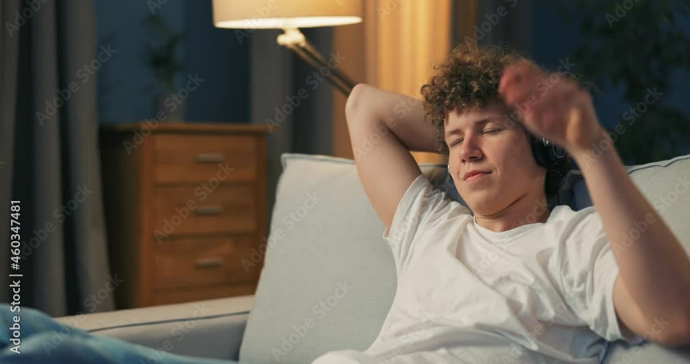 A young man is resting on a sofa, hands behind head, turns off tv listening to music on wireless headphones on ears, eyes closed, listening to a song, immersed in a dream, nodding, smiling singing A young man is resting on a sofa, hands behind head, turns off tv listening to music on wireless headphones on ears, eyes closed, listening to a song, immersed in a dream, nodding, smiling singing