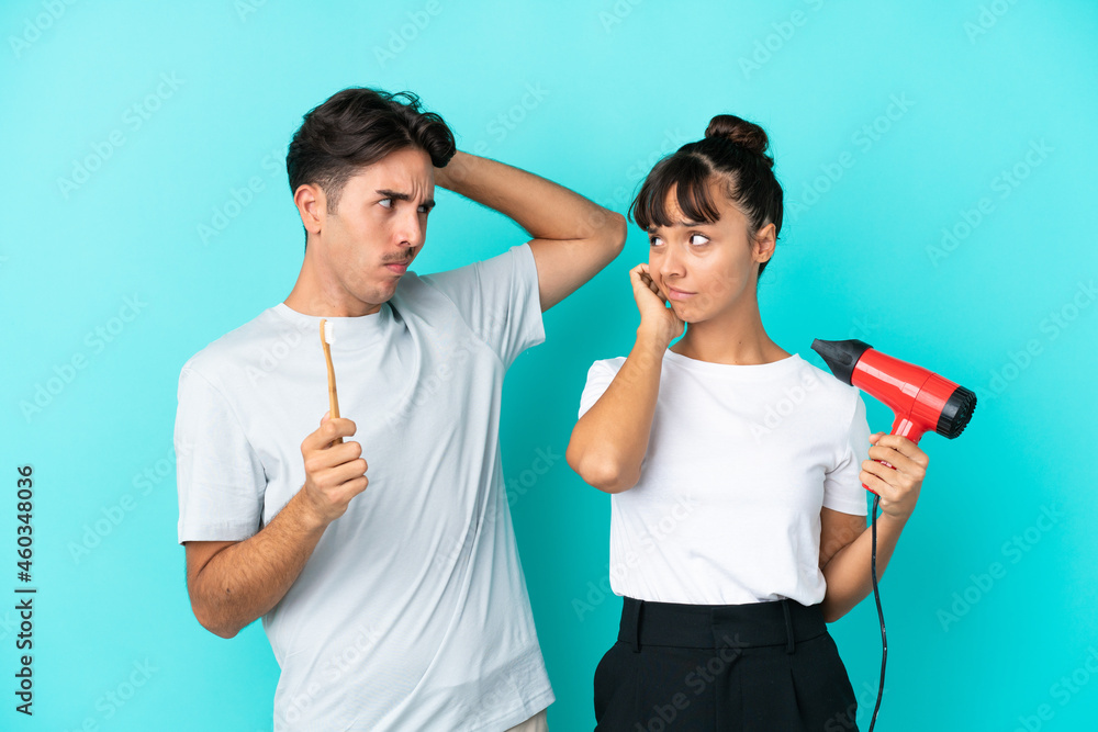 Young mixed race couple holding a hairdryer and toothbrush isolated on blue background having doubts while scratching head Young mixed race couple holding a hairdryer and toothbrush isolated on blue background having doubts while scratching head