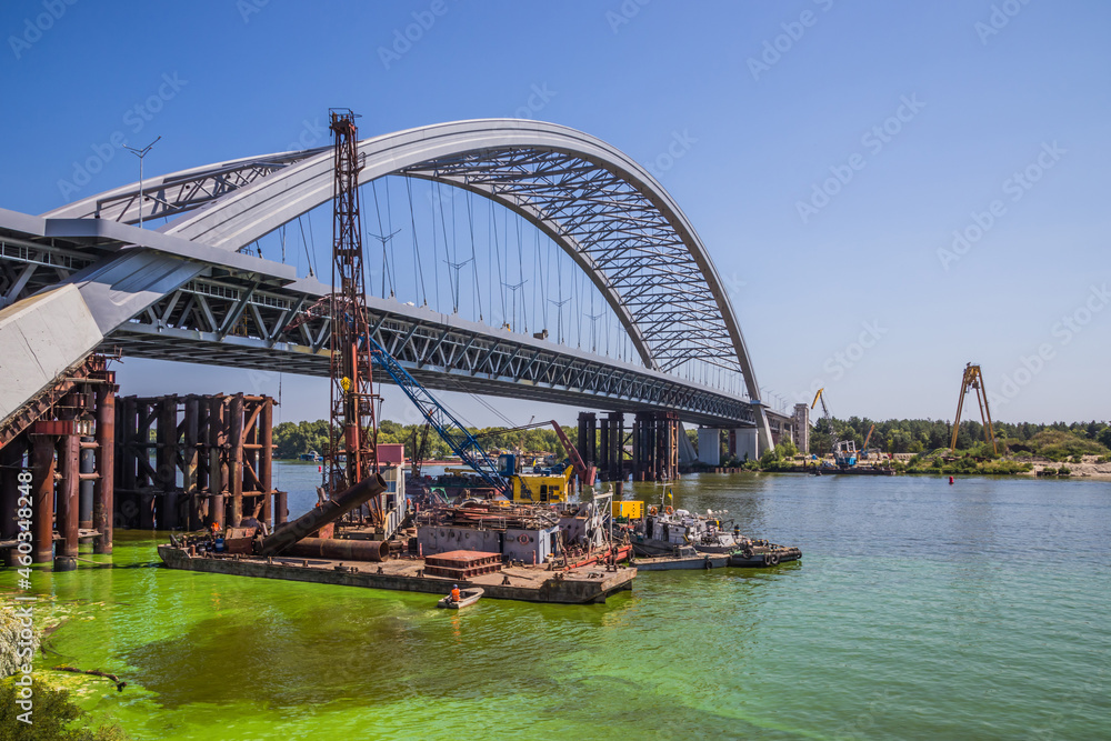 Fototapeta premium Construction of a large arched bridge in the capital of Ukraine. Podolsky bridge, panoramic view of the bridge under construction across the Dnipro, clear weather, summer. Kyiv