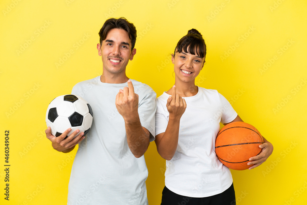 young sport couple playing football and basketball isolated on yellow background inviting to come with hand. Happy that you came