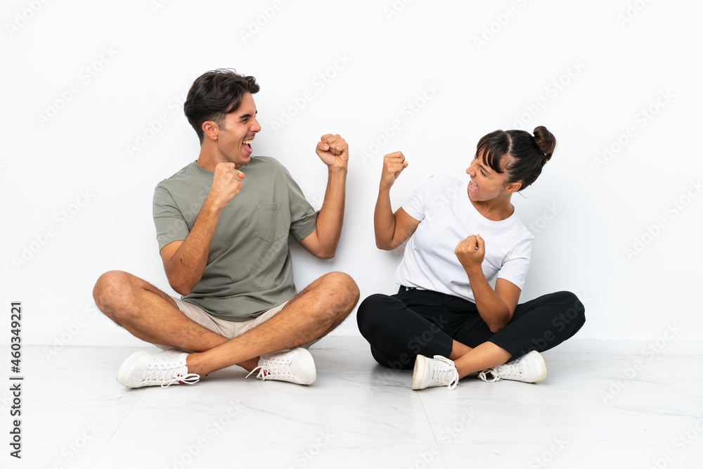 Young mixed race couple sitting on the floor isolated on white background celebrating a victory in winner position Young mixed race couple sitting on the floor isolated on white background celebrating a victory in winner position