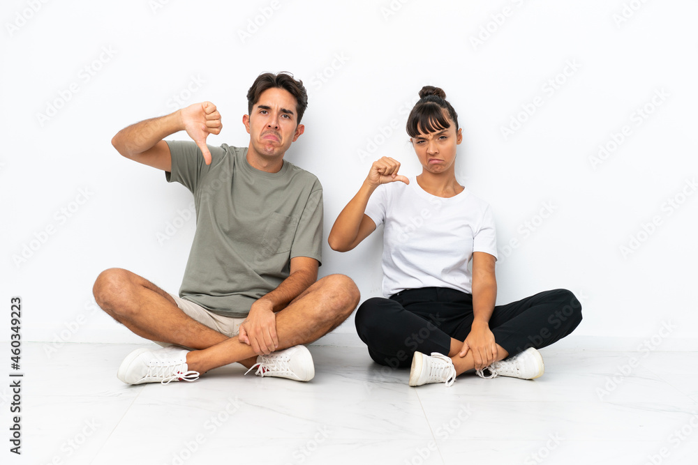 Young mixed race couple sitting on the floor isolated on white background showing thumb down sign with negative expression Young mixed race couple sitting on the floor isolated on white background showing thumb down sign with negative expression