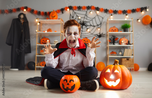 Boo, scare on Halloween. Portrait of terrifying little boy in traditional Halloween vampire costume who screams and scares looking at camera. Child sits on floor near pumpkin in room with festive deor photo