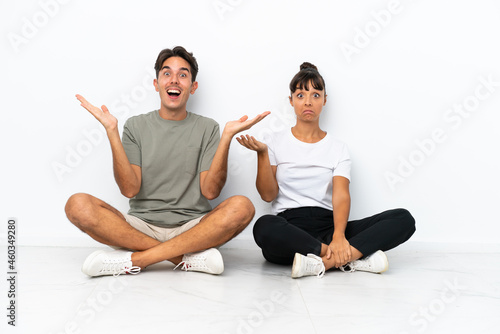 Young mixed race couple sitting on the floor isolated on white background holding copyspace imaginary on the palm to insert an ad