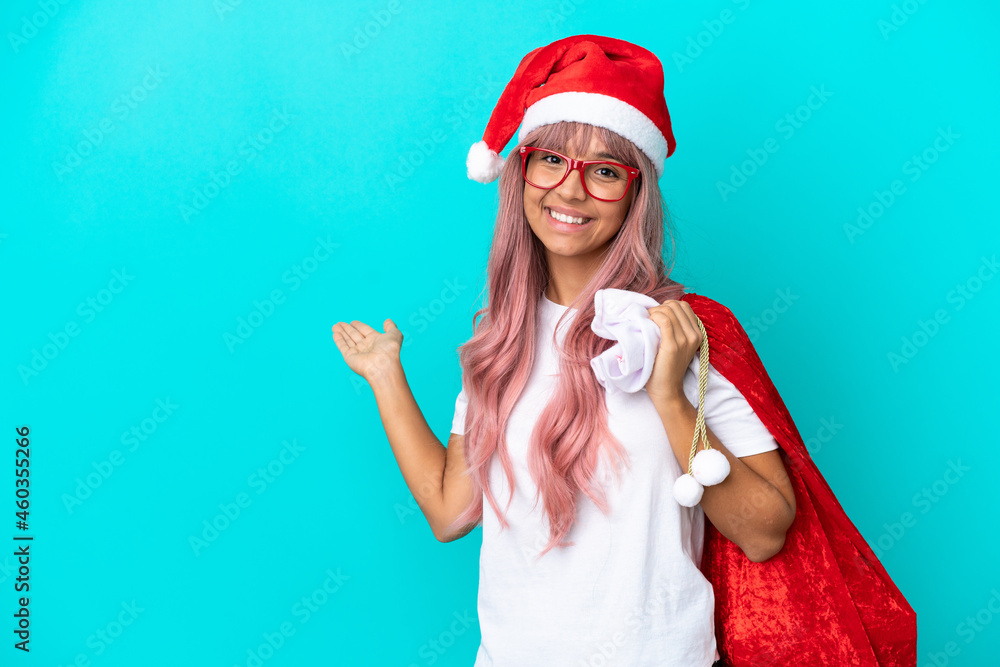 Young mixed race woman handing out gifts on Christmas isolated on blue background extending hands to the side for inviting to come Young mixed race woman handing out gifts on Christmas isolated on blue background extending hands to the side for inviting to come
