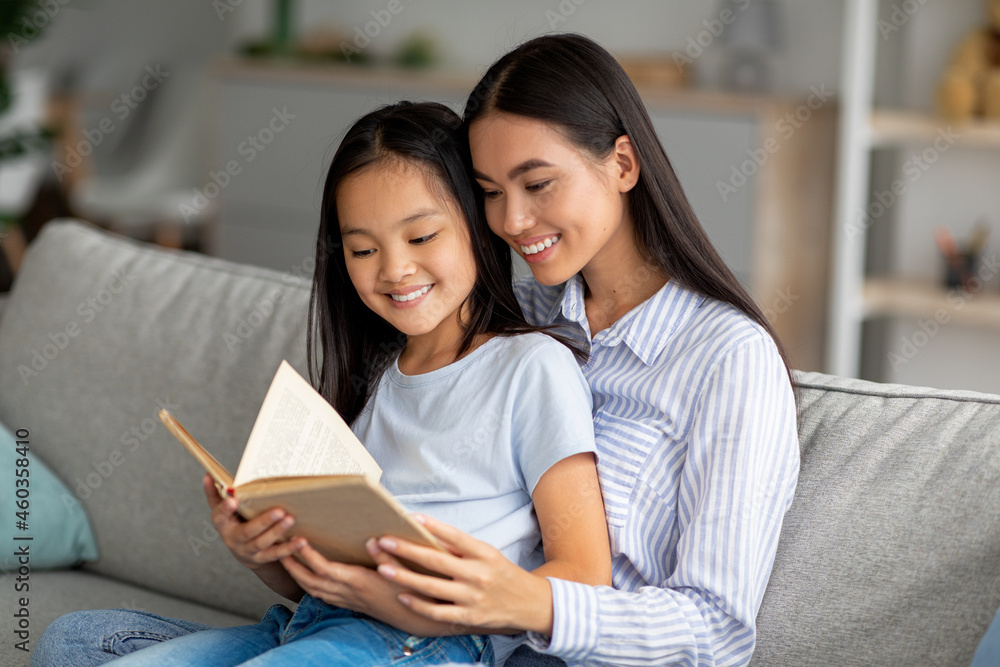 Loving young asian mother reading book with her pretty daughter, sitting together on sofa at home Loving young asian mother reading book with her pretty daughter, sitting together on sofa at home