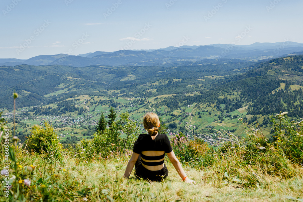 Naklejka premium Little girl in a black T-shirt sitting on the green green meadow in Caprathian mountains. Back view. Travel, enjoying life, admire the beautiful nature view.