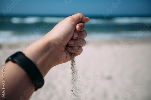 Sand is pouring out of a woman's hand against the background of the ocean, blue sky and white sand on the beach. Big waves on the sea. There is a black bracelet on her hand. Close-up photo