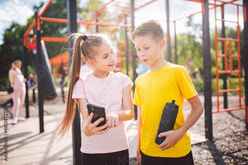 Technology and sport for children. Two teenage kids using smartphone and online workout app. Twins boy and girl watching video on phone during break from sports lesson at school at outside gym