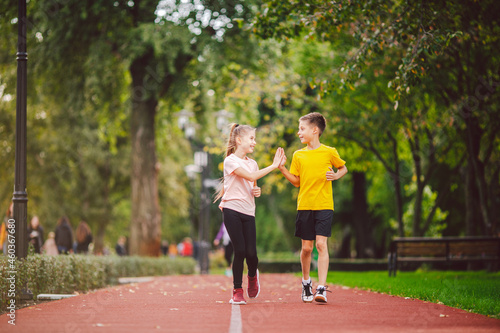 Fototapeta Naklejka Na Ścianę i Meble -  Sports and fitness in adolescence. Caucasian twins boy and girl run on the jogging track in the city park. Two children brother and sister for 10 years running on a rubberized outdoor treadmill