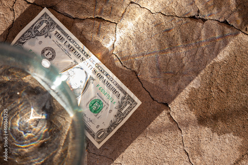 An overhead view of a jar of water and a dollar lying on a cracked concrete pavement in a beam of refracted and split sunlight. Selective focus.