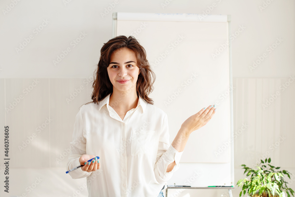 Portrait of young woman posing near blank empty whiteboard. Presentation concept. Teacher in white, businesswoman standing with a marker near a whiteboard, Online classes, business training Portrait of young woman posing near blank empty whiteboard. Presentation concept. Teacher in white, businesswoman standing with a marker near a whiteboard, Online classes, business training