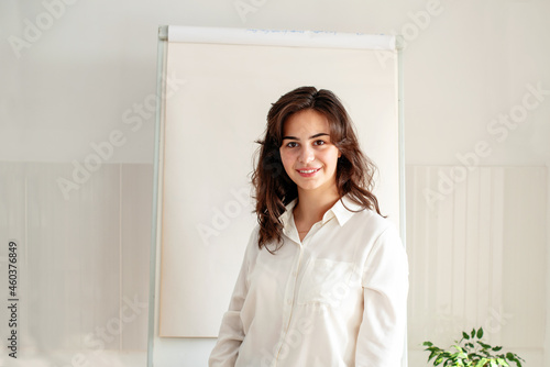 Portrait of young woman posing near blank empty whiteboard. Presentation concept. Teacher in white  businesswoman standing with a marker near a whiteboard  Online classes  business training 