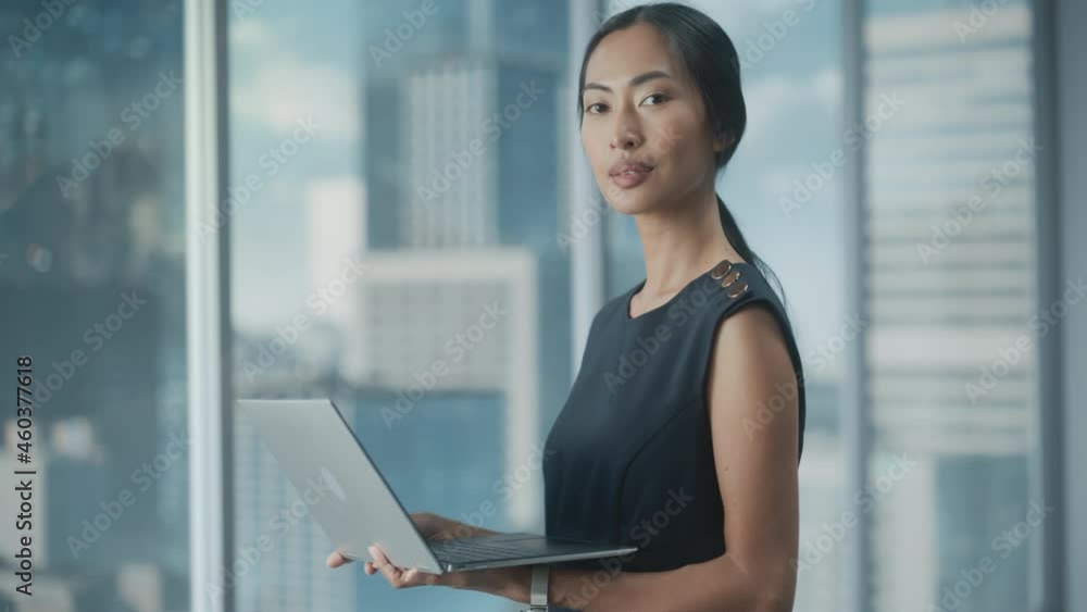 Beautiful Portrait of an Asian Businesswoman in Stylish Black Dress Using Laptop Computer, Posing Next to Window in City Office. Confident Female CEO Smiling. Successful Diverse Business Manager. Beautiful Portrait of an Asian Businesswoman in Stylish Black Dress Using Laptop Computer, Posing Next to Window in City Office. Confident Female CEO Smiling. Successful Diverse Business Manager.