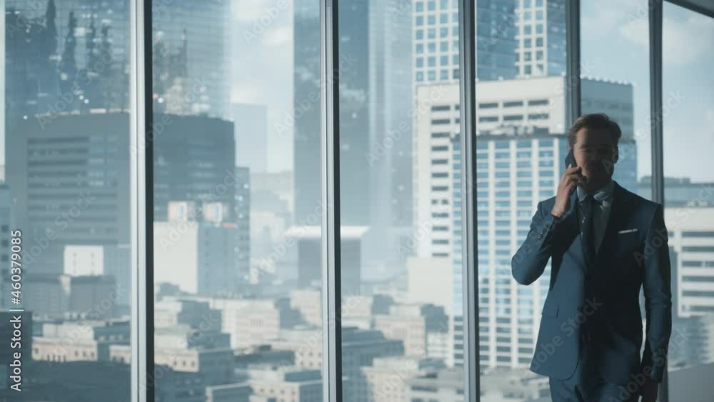 Confident Young Businessman in a Suit Walking in Modern Office, Talking on a Phone, Looking out of the Window on Big City with Skyscrapers. Successful Finance Manager Planning Work Projects. Confident Young Businessman in a Suit Walking in Modern Office, Talking on a Phone, Looking out of the Window on Big City with Skyscrapers. Successful Finance Manager Planning Work Projects.