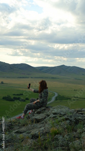 Young woman with curly hair is sitting in camouflage overalls at the top of the mountain. Sit with your back. The woman takes a photo. Mountain top, mountain landscape. Tourist hike up the mountain. 