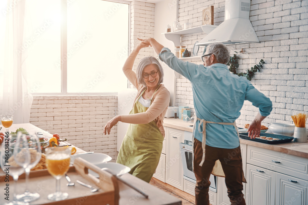 Beautiful playful senior couple in aprons dancing and smiling while preparing healthy dinner at home - obrazy, fototapety, plakaty 