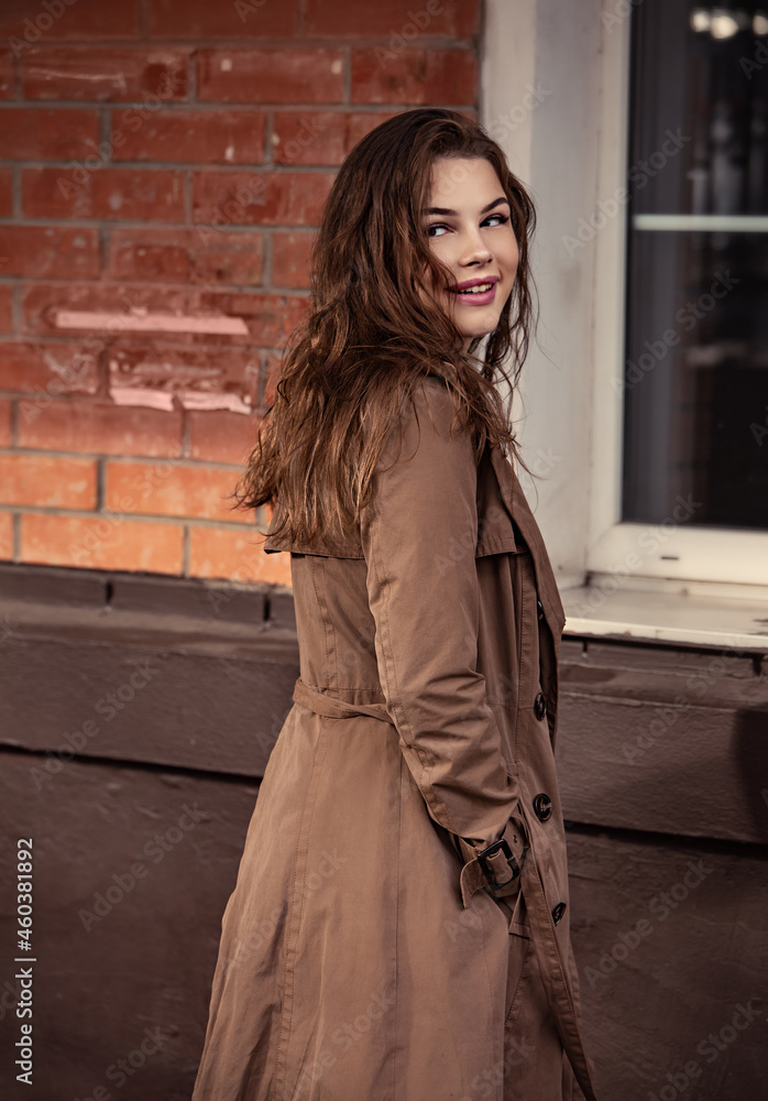 Fototapeta premium Young smiling woman walking on the street city wall background in brown coat and black shoes near the old bulding with big windows. Autumn