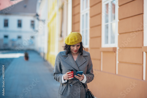 Woman using smartphone and drinking coffee takeaway while wearing yellow beret and a gray coat