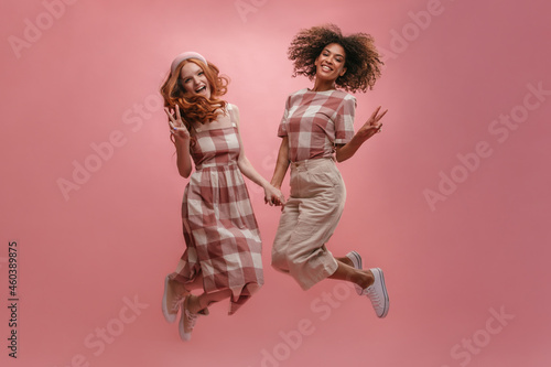 Jumping young girls of different race in linen gingham clothes showing peace sign on pink background. White girl with foxy hair in summer dress and black girl with dark volume curls in light dress.
