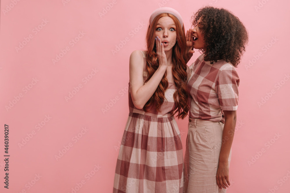 African girl with dark curls tells exciting news to her red-haired caucasian friend. White slender girl in summer pink dress is surprised at what she hears. Emotions of people, lifestyle concept. African girl with dark curls tells exciting news to her red-haired caucasian friend. White slender girl in summer pink dress is surprised at what she hears. Emotions of people, lifestyle concept.