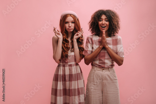 Attractive caucasian lady crosses her fingers, african woman holds palms together on pink background. Beauties, brunette and redhead are dressed in summer plaid casual clothes in hope of good news.