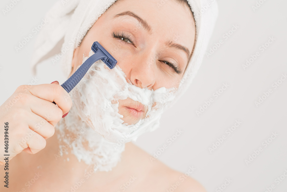Cheerful caucasian woman with a towel on her head and shaving foam on her face holds a razor on a white background