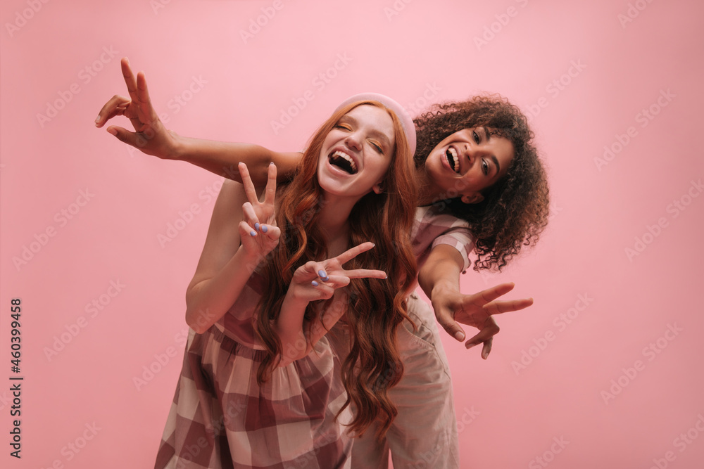 International young girls in good mood showing peace sign on pink background. African and caucasian ladies with happy wide smiles wear clothes in different shades. People sincere emotions concept. International young girls in good mood showing peace sign on pink background. African and caucasian ladies with happy wide smiles wear clothes in different shades. People sincere emotions concept.
