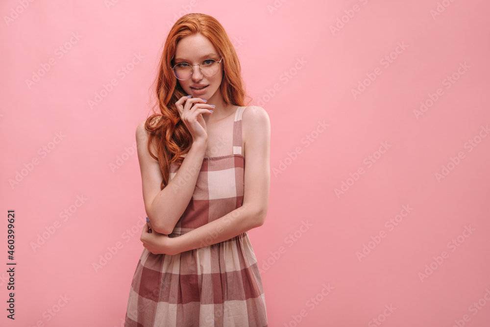 Red-haired caucasian girl sly look at camera with copy space. Fox, like lady in different shades of pink sundress in cage and in big glasses, holding her hand near chin. Red-haired caucasian girl sly look at camera with copy space. Fox, like lady in different shades of pink sundress in cage and in big glasses, holding her hand near chin.