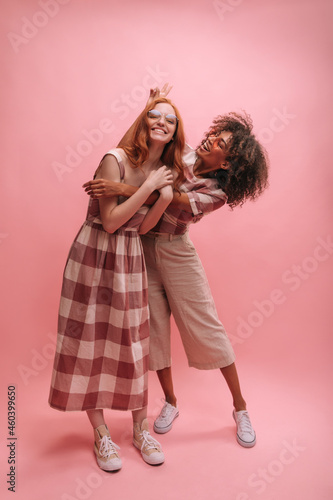 Full length image of stylish interracial girlfriends fooling around in studio with copy space. African brunette girl puts bunny ears over head of caucasian redhead teenager.